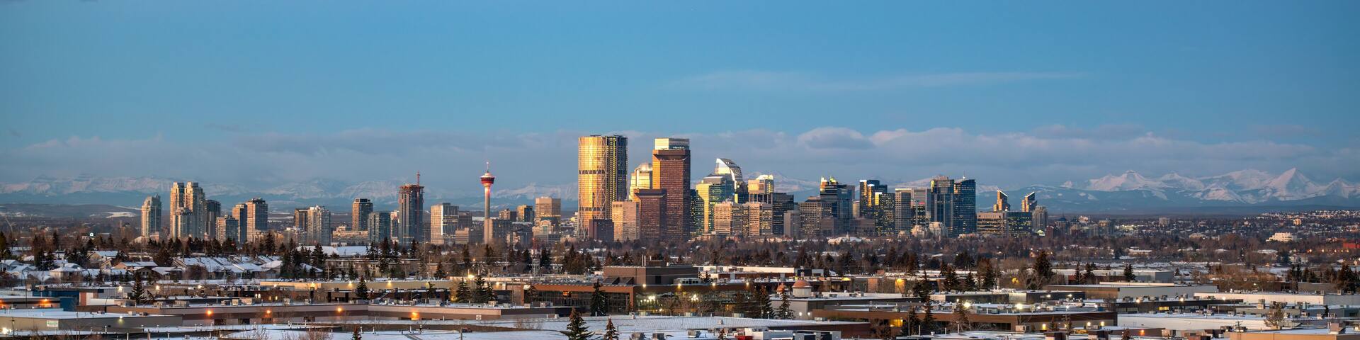A city skyline with modern buildings and a tower, surrounded by snow-covered trees and mountains in the background.