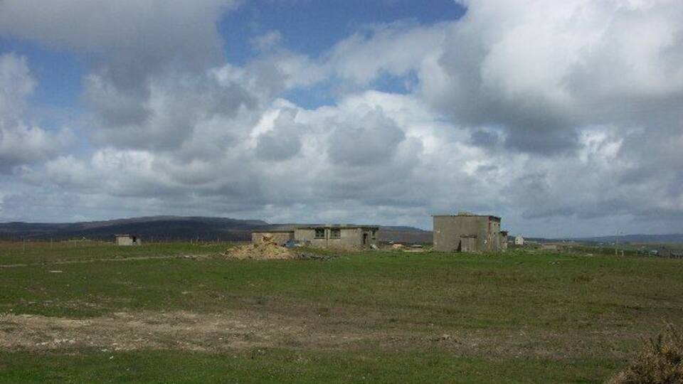 A grassy field with two small, dilapidated buildings and a hill in the background.