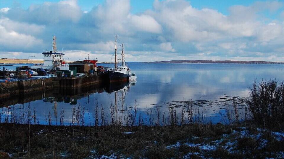 A calm harbor with a docked ship, a lighthouse, and a snow-covered shoreline.
