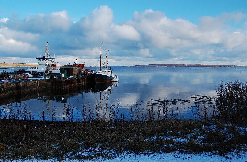 A calm harbor with a docked ship, a lighthouse, and a snow-covered shoreline.