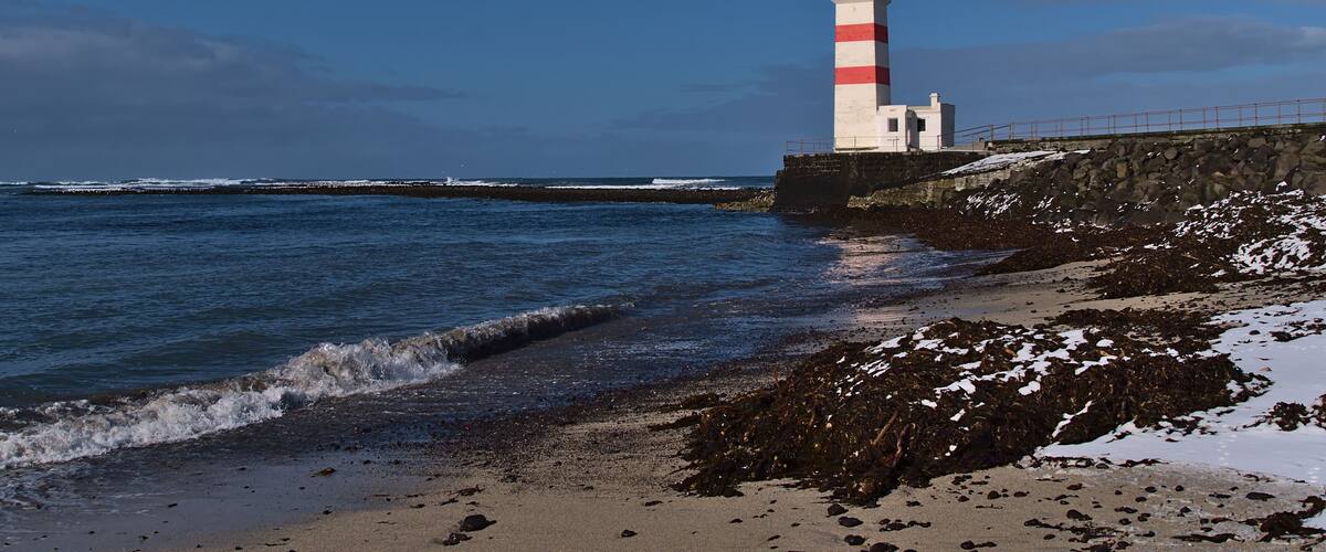 Beautiful view of white and red colored old lighthouse Garður on the coast of Suðurnesjabær on Reykjanes peninsula, Iceland with beach and snow in winter season on sunny day.