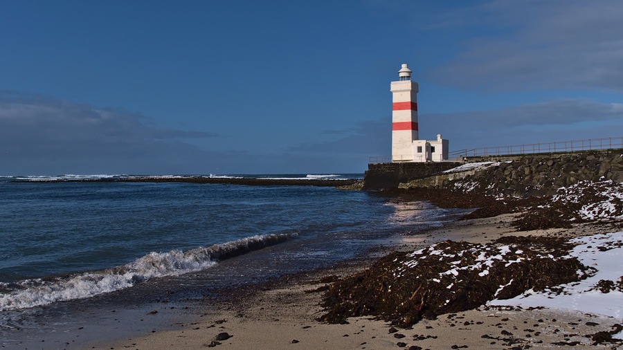 Beautiful view of white and red colored old lighthouse Garður on the coast of Suðurnesjabær on Reykjanes peninsula, Iceland with beach and snow in winter season on sunny day.