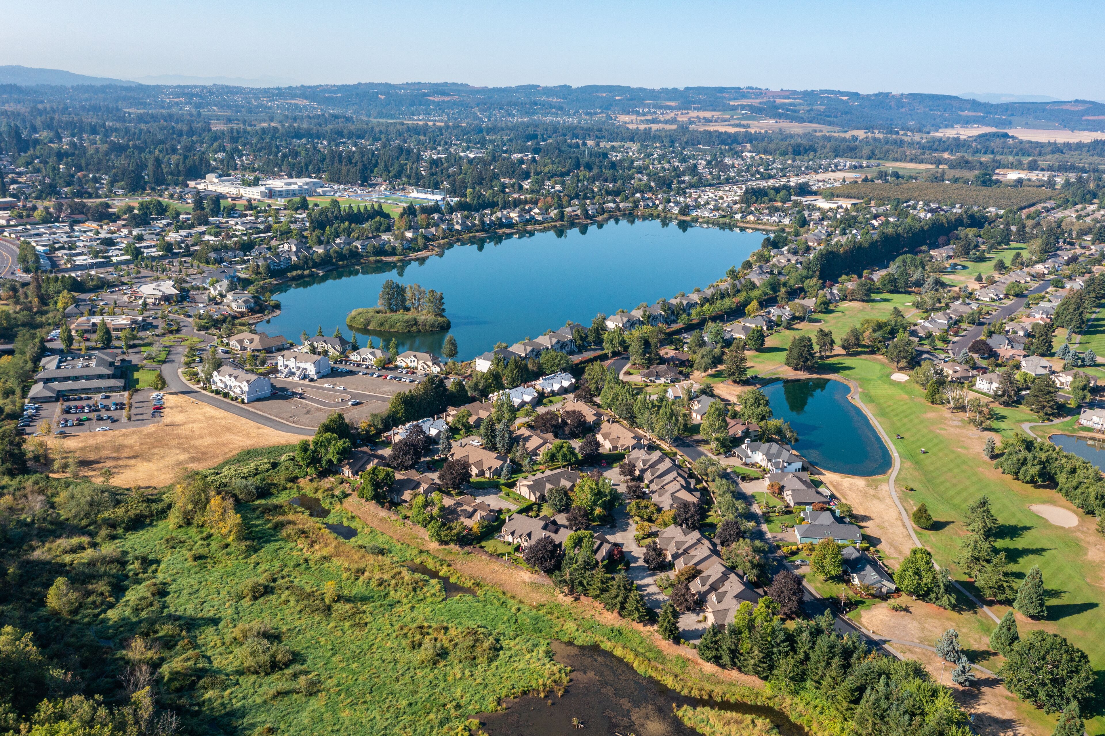 Aerial drone view of Staats Lake in Keizer, Oregon, near Salem, featuring a residential neighborhood, a golf course, and lush green landscapes under a clear blue sky