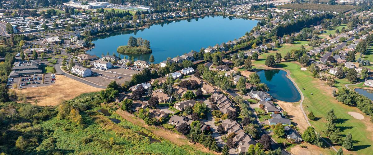 Aerial drone view of Staats Lake in Keizer, Oregon, near Salem, featuring a residential neighborhood, a golf course, and lush green landscapes under a clear blue sky