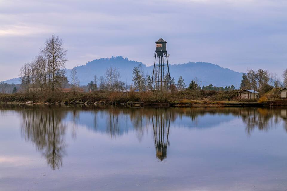 Ein ruhiger See mit einem Wasserturm, Bäumen und Bergen im Hintergrund.