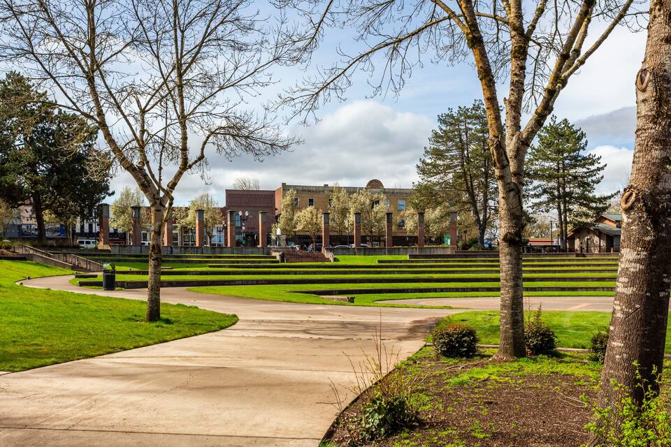 A landscaped area with a concrete pathway, trees, and a building in the background.