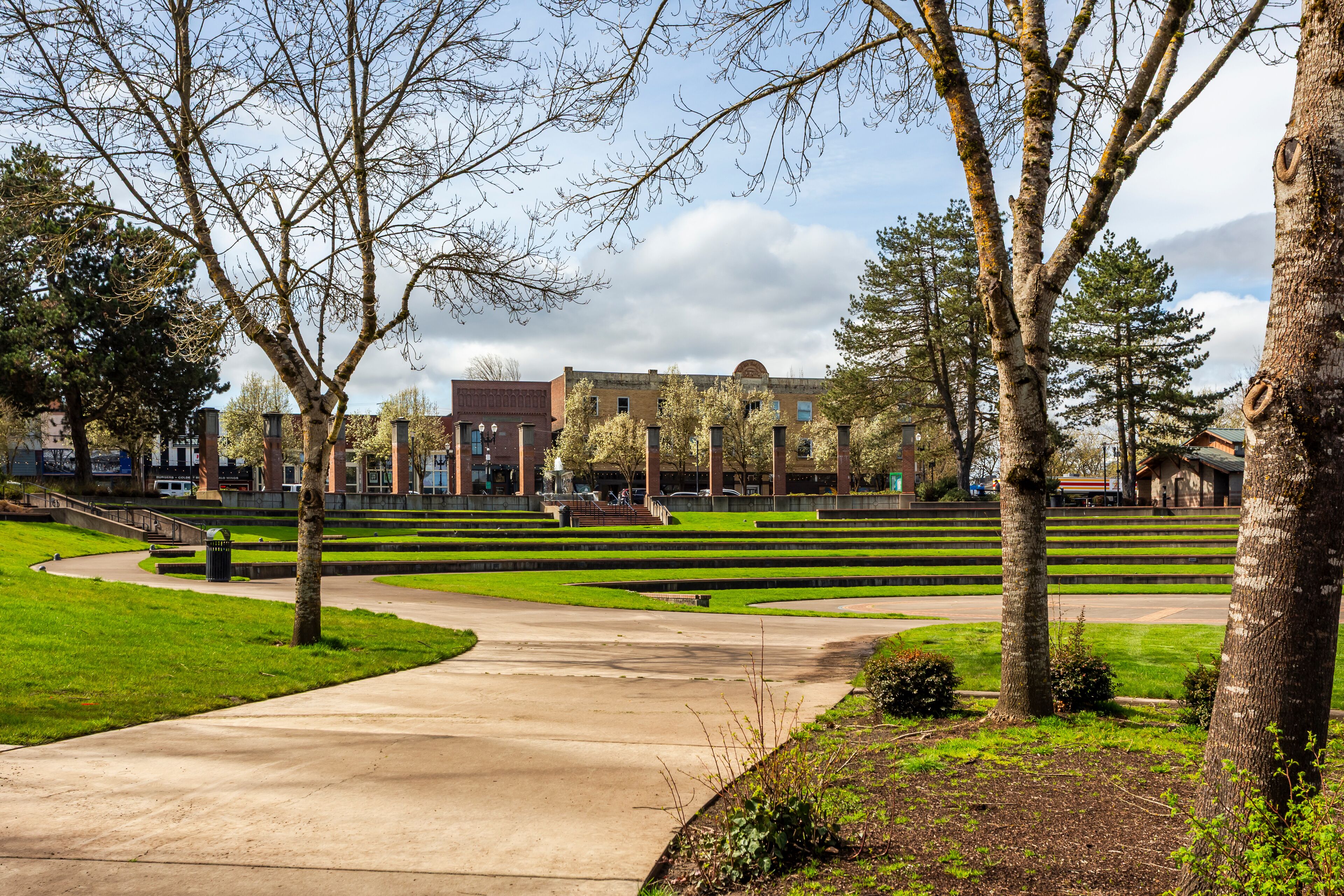A landscaped area with a concrete pathway, trees, and a building in the background.