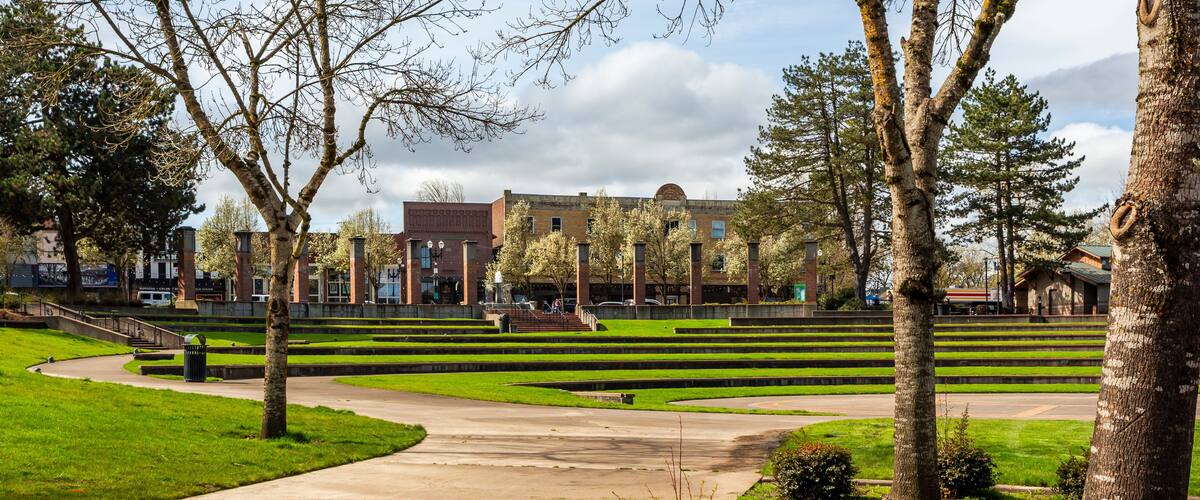 A landscaped area with a concrete pathway, trees, and a building in the background.