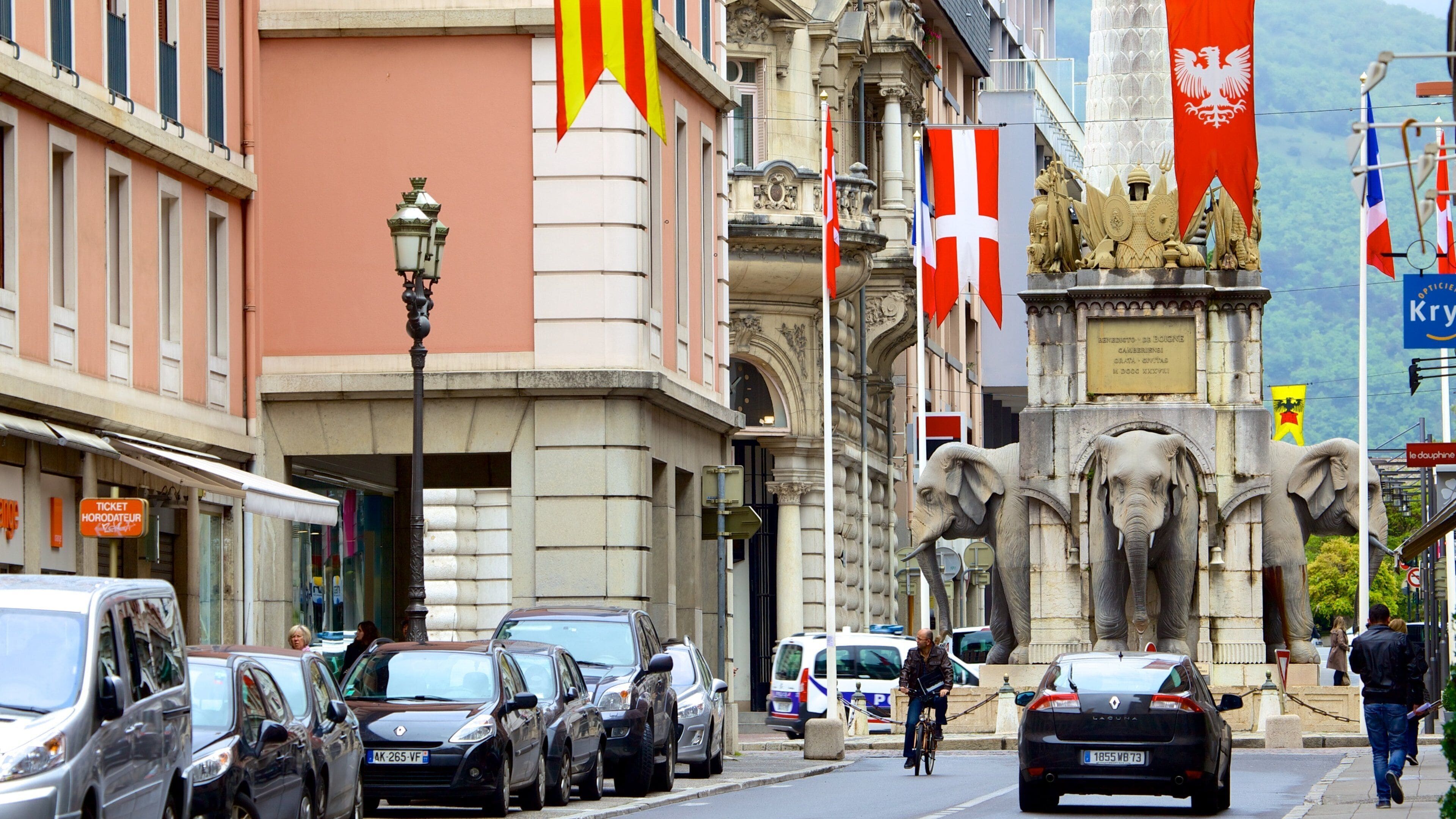 A street with parked cars, a man on a bicycle, and a statue of elephants. Flags of various countries are visible.