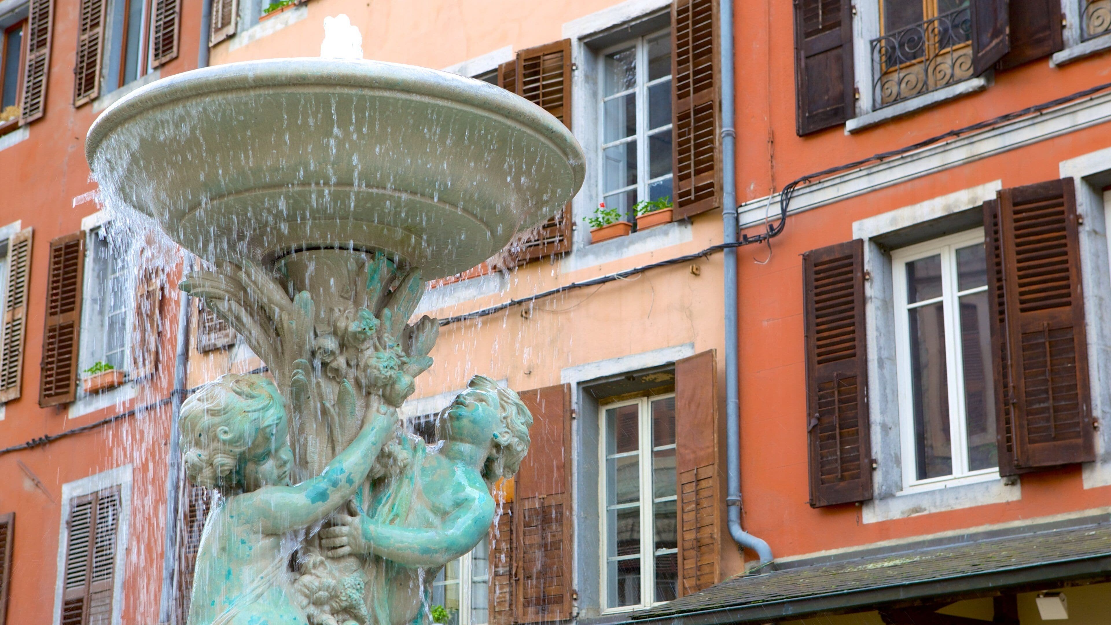 A fountain with water spouting from a large basin, surrounded by buildings with orange facades and brown shutters.