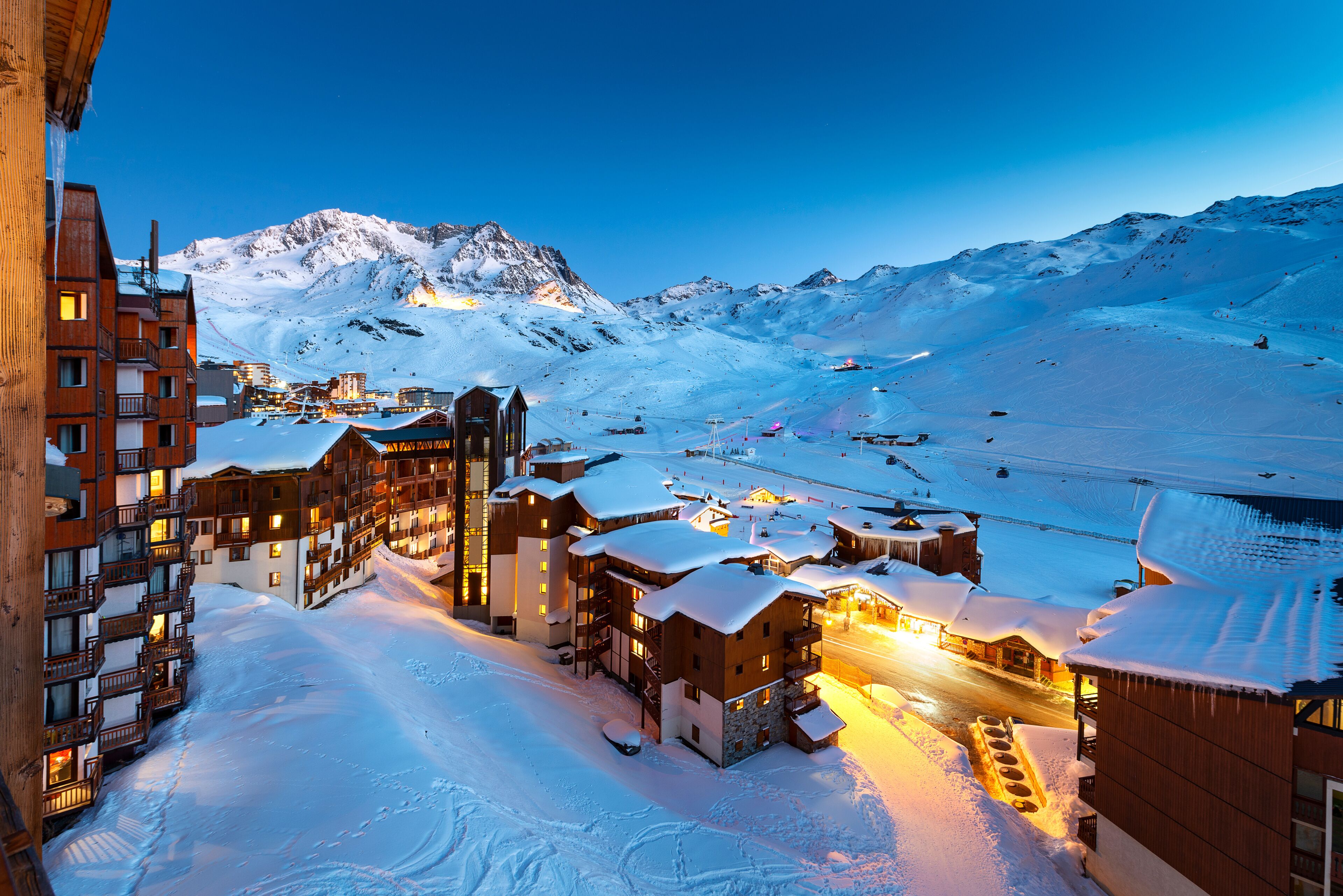 A snow-covered mountain town at dusk with illuminated buildings and ski slopes.