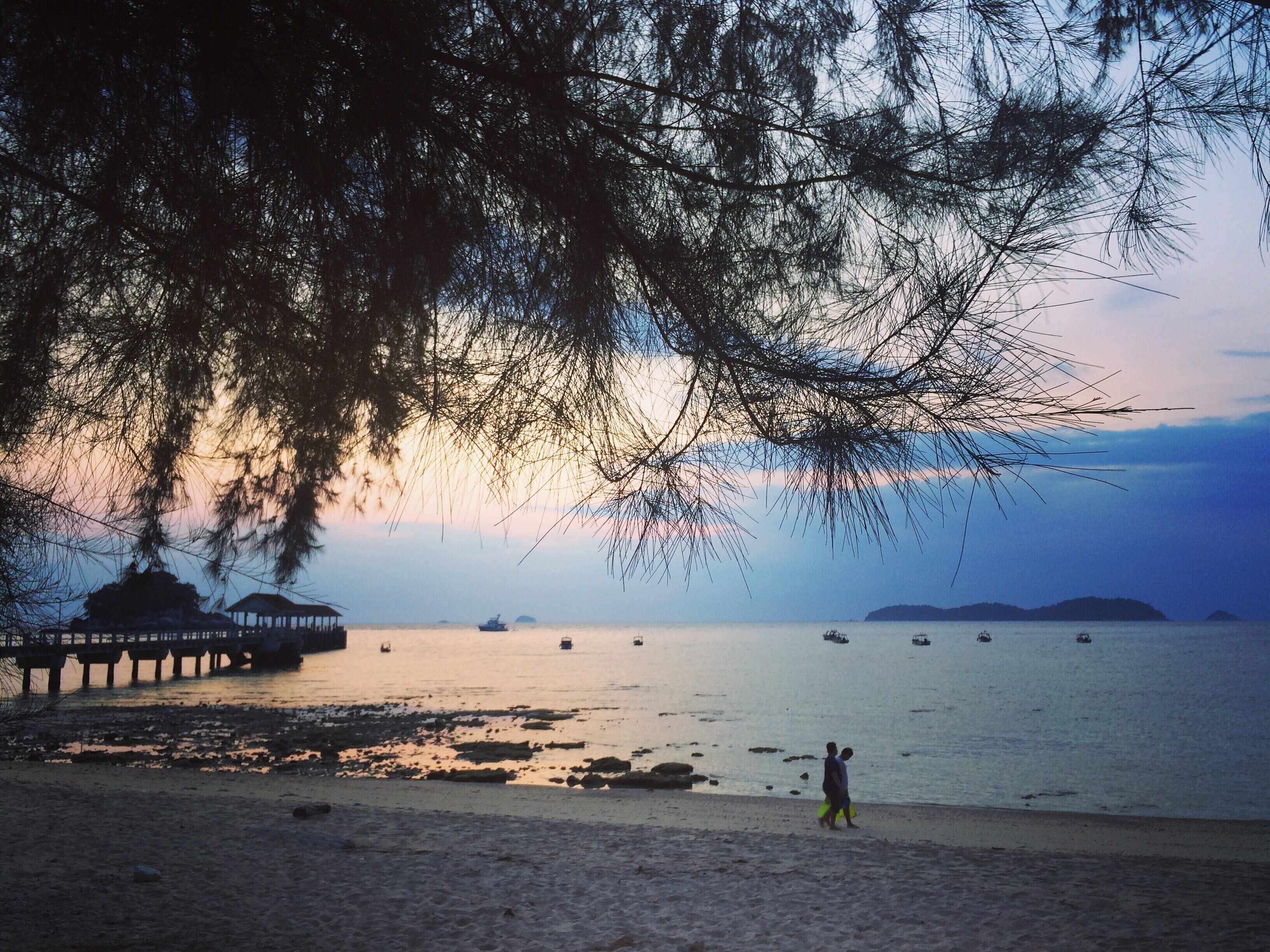 A beach with a pier and boats in the distance during sunset.