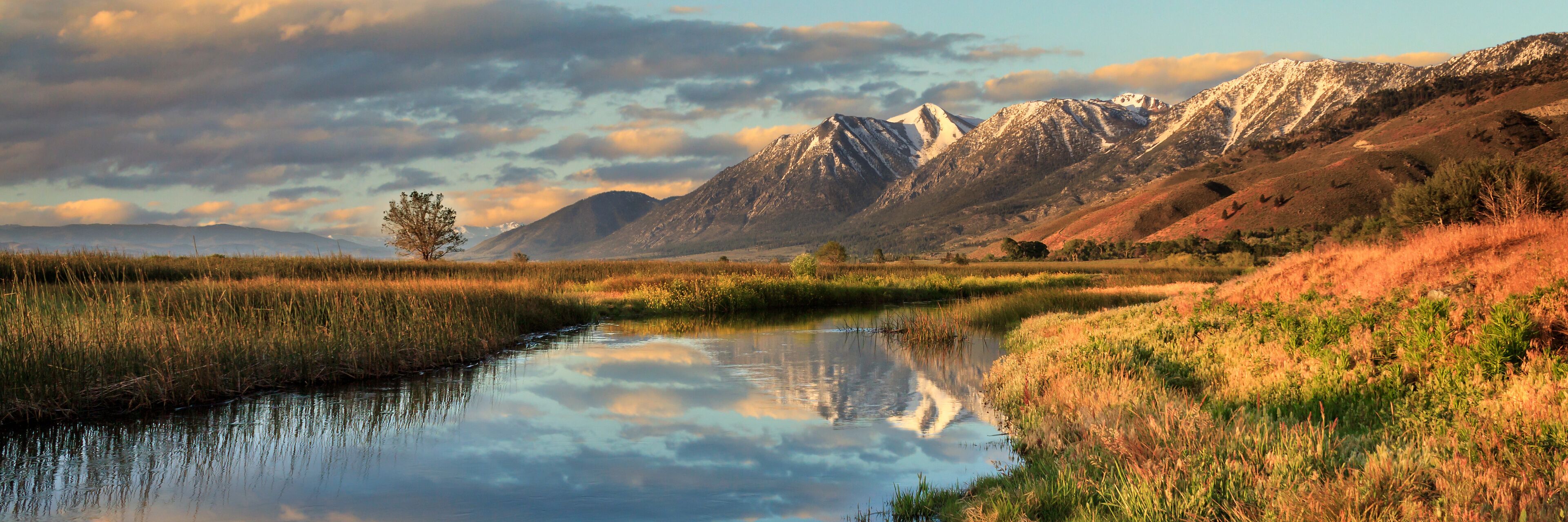 A calm river reflecting the surrounding landscape, with mountains in the background and a clear sky.