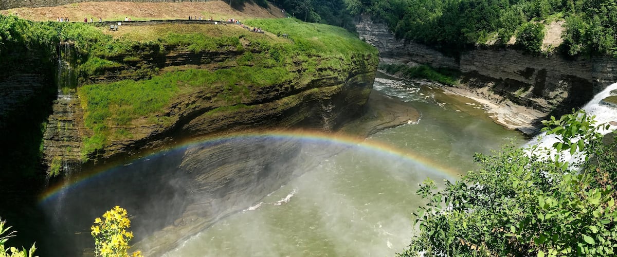 A river with a rainbow, green hills, and a stone wall.