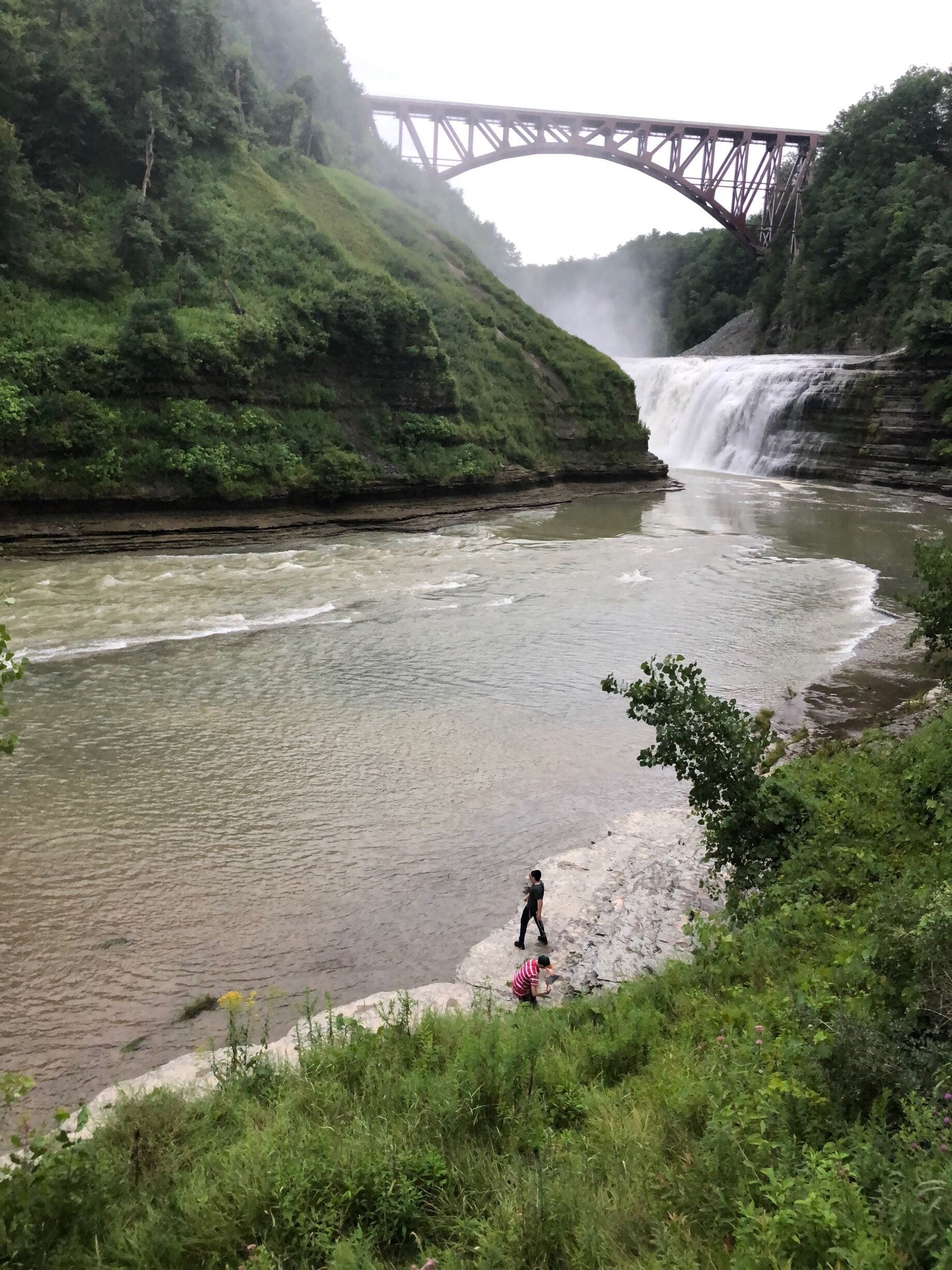A river with a waterfall, a bridge, and a person walking near the water.