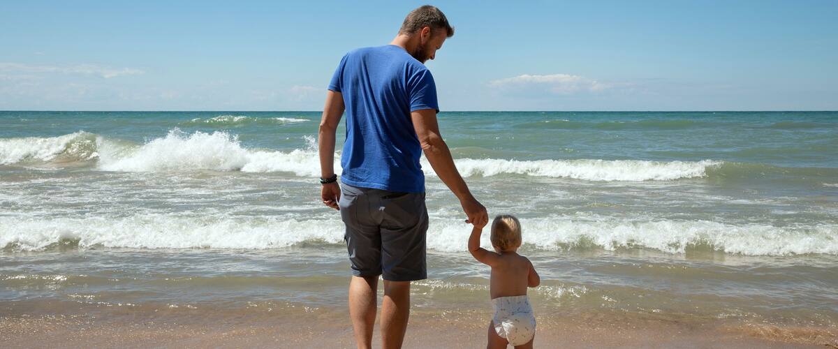 A man and a child holding hands on a beach with waves in the background.