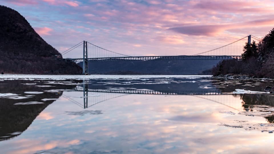 A suspension bridge over a calm river with reflections, surrounded by hills and a clear sky.
