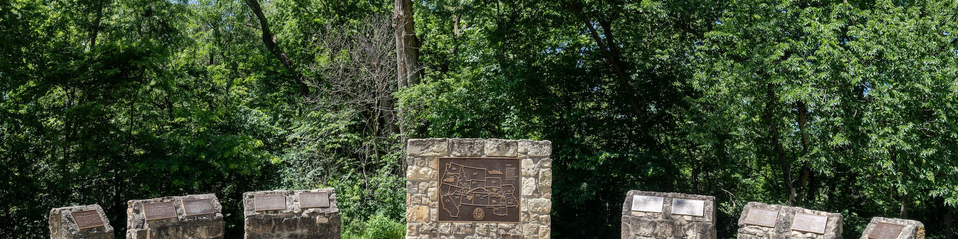 A stone monument with inscriptions, surrounded by a paved area and dense green trees.