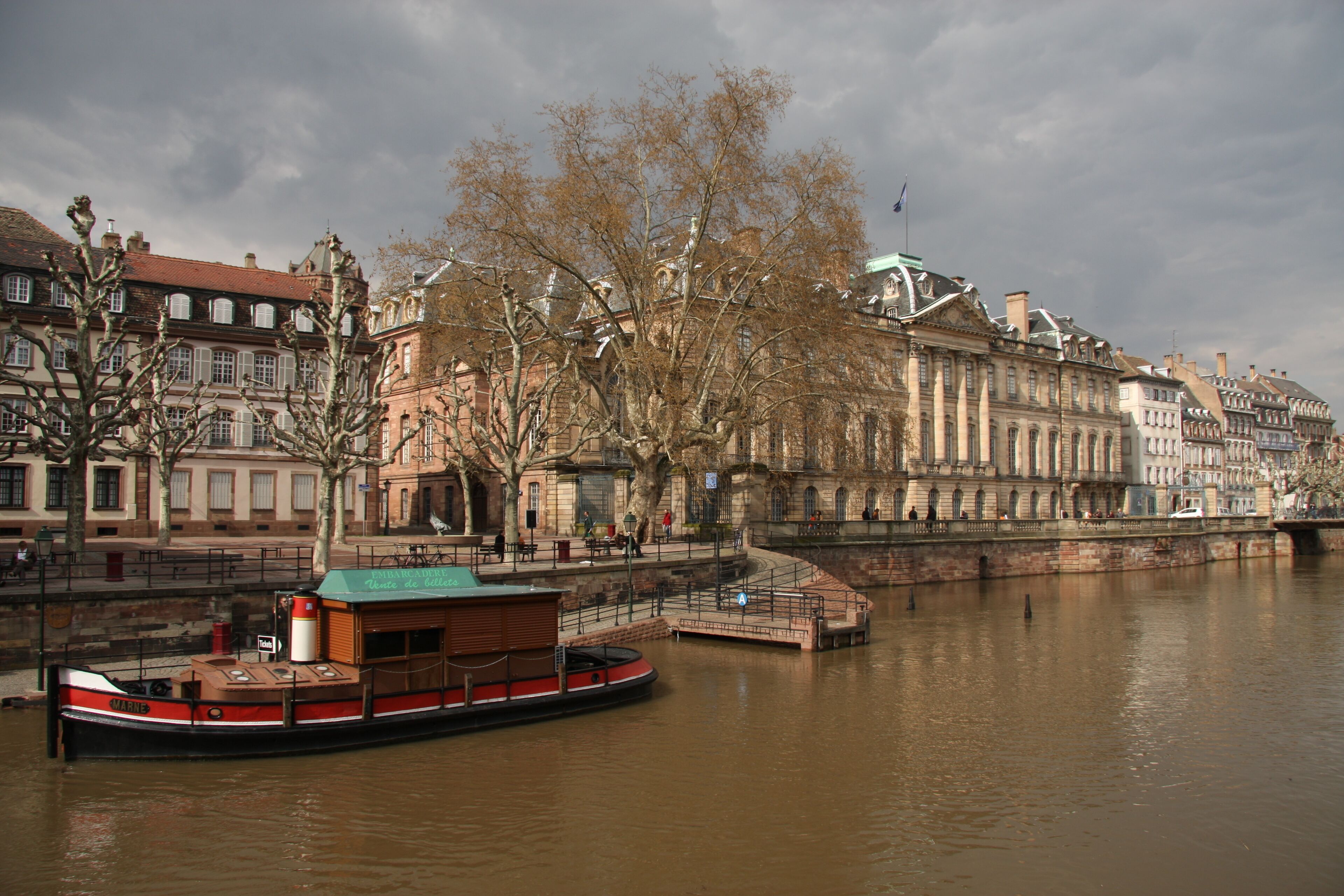 A river with a boat, buildings, and leafless trees.