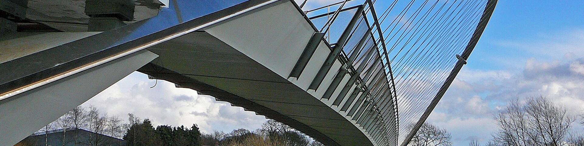 A modern bridge with a curved design over a river, surrounded by greenery and buildings.