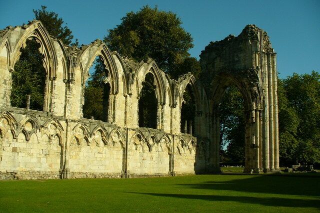 A Gothic-style stone structure with multiple arches and columns, surrounded by a grassy area and trees.