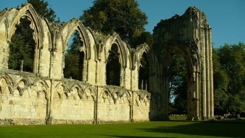 A Gothic-style stone structure with multiple arches and columns, surrounded by a grassy area and trees.