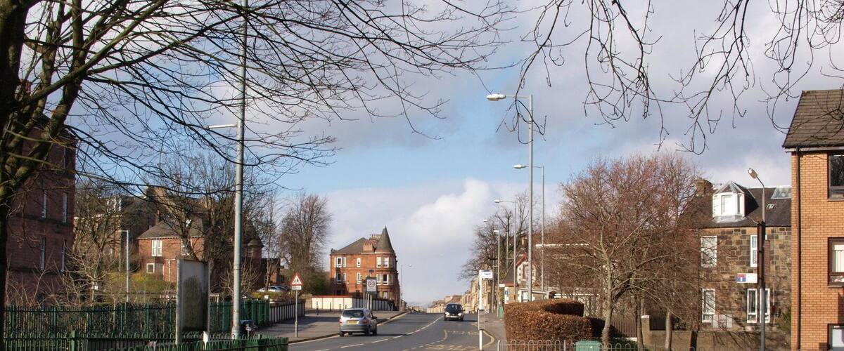 A quiet residential street with houses, a green fence, and a clear sky.