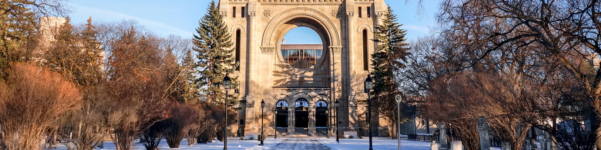 A snow-covered pathway leading to a large stone archway with a statue on top, surrounded by leafless trees and a clear sky.