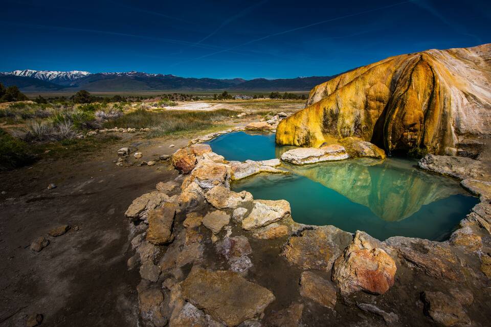 Una fuente termal geotérmica con depósitos minerales, rodeada de un paisaje montañoso.
