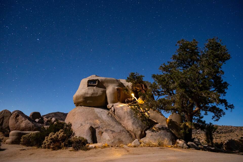 A rock formation with a cave-like structure and a tree under a starry night sky.
