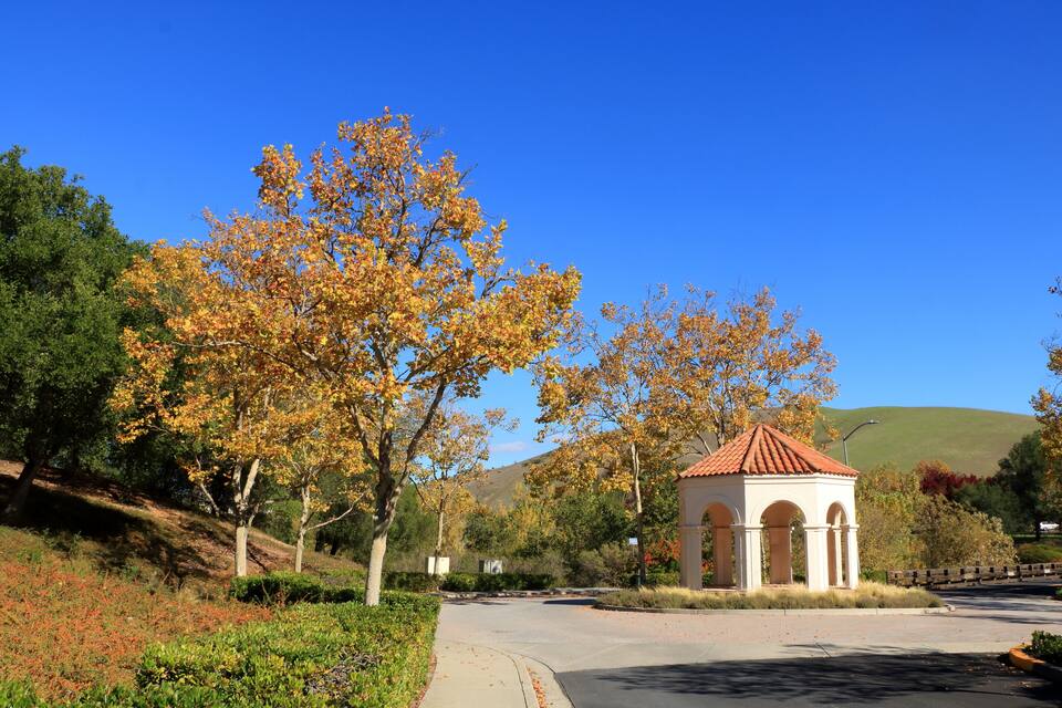 A gazebo with a red roof surrounded by trees with autumn foliage, a clear blue sky, and a hilly landscape in the background.