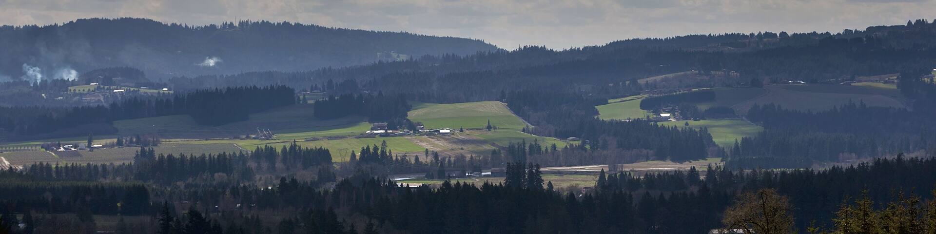 A landscape with rolling hills, scattered trees, and a partly cloudy sky.