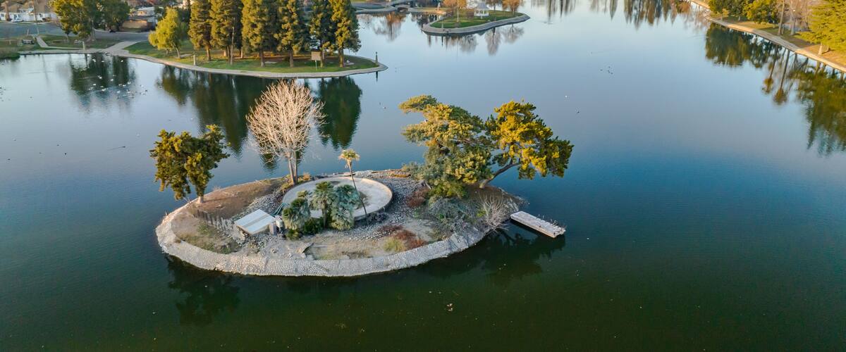 A tranquil lake with a small island featuring a gazebo, surrounded by dense trees and a distant urban landscape.