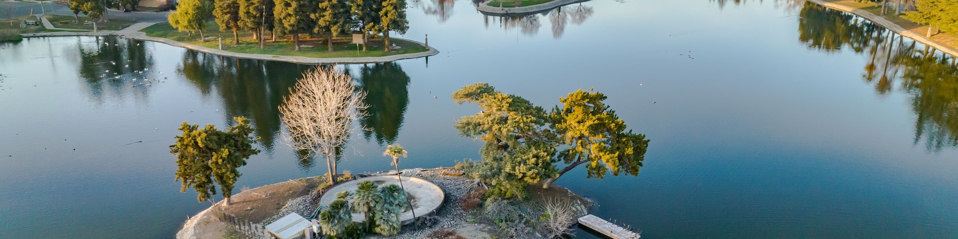A tranquil lake with a small island featuring a gazebo, surrounded by dense trees and a distant urban landscape.