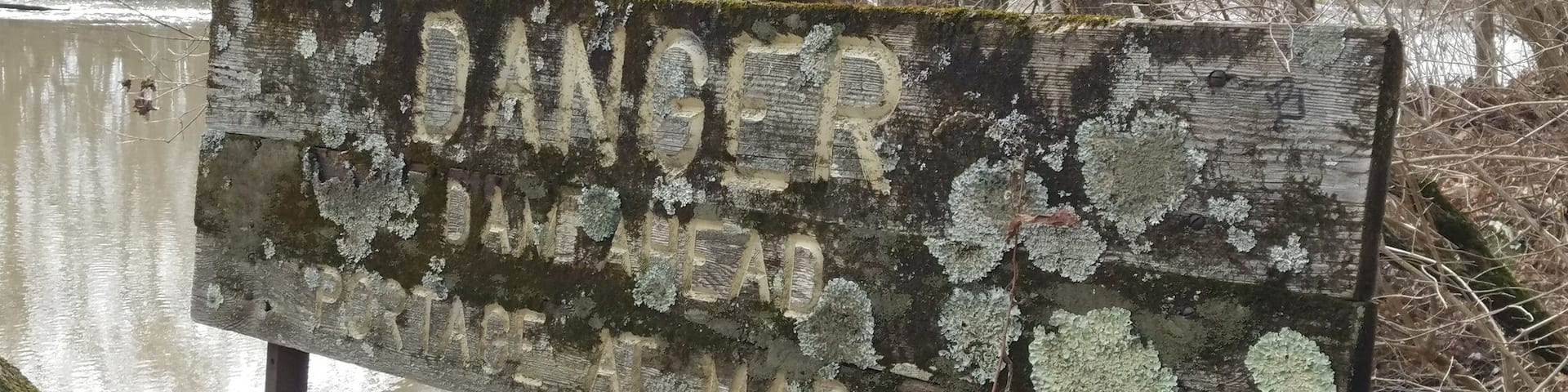An old wooden sign warning about the lowhead dam just down stream. There are newer looking metal signs. The lichen encrusted sign reads "Danger, Dam Ahead. Portage at Marker". This can be found east of Antrim lake on the western shore of the river, along the trails that snake through the wooded area of the park. #urbanjungle