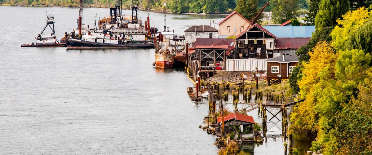 A river with a large industrial ship docked, surrounded by wooden buildings and a forested hillside.