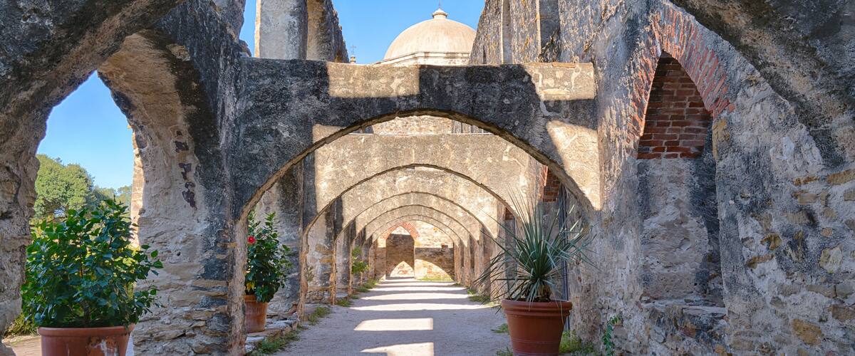 An ancient stone corridor with arches, potted plants, and a dome structure in the background.