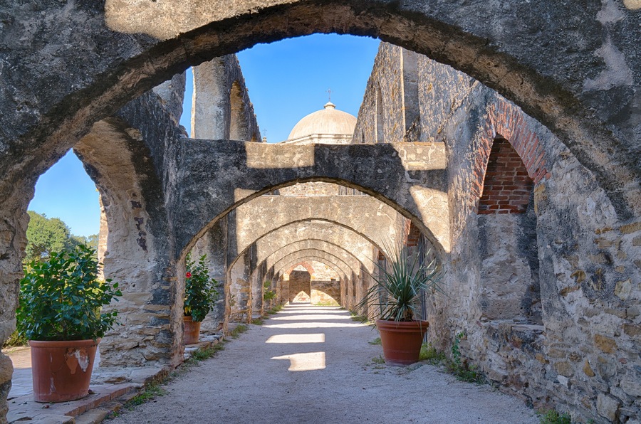 Un antiguo corredor de piedra con arcos, plantas en macetas y una estructura abovedada a lo lejos.