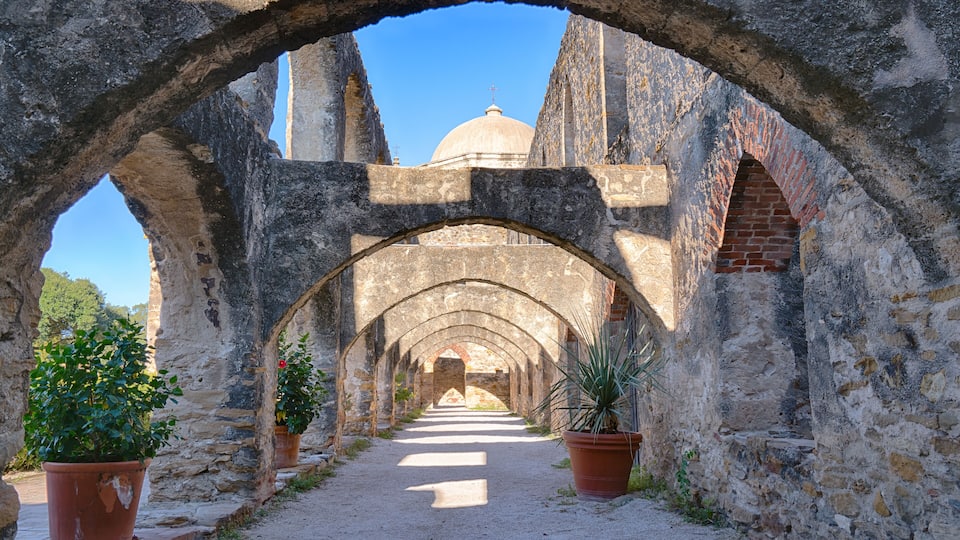 An ancient stone corridor with arches, potted plants, and a distant dome structure.