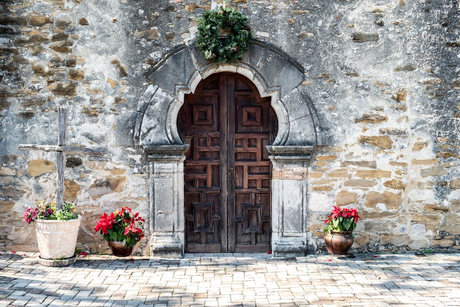Un edificio de piedra con una gran puerta doble de madera, dos macetas con flores rojas y una guirnalda sobre la puerta.