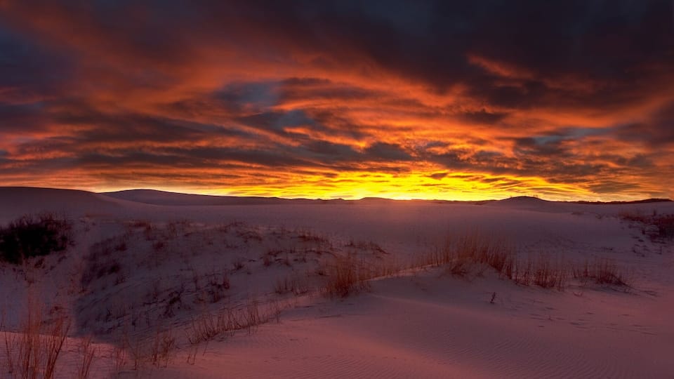 A desert landscape with rolling sand dunes and a dramatic sunset.