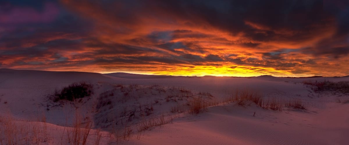 A desert landscape with rolling sand dunes and a dramatic sunset.