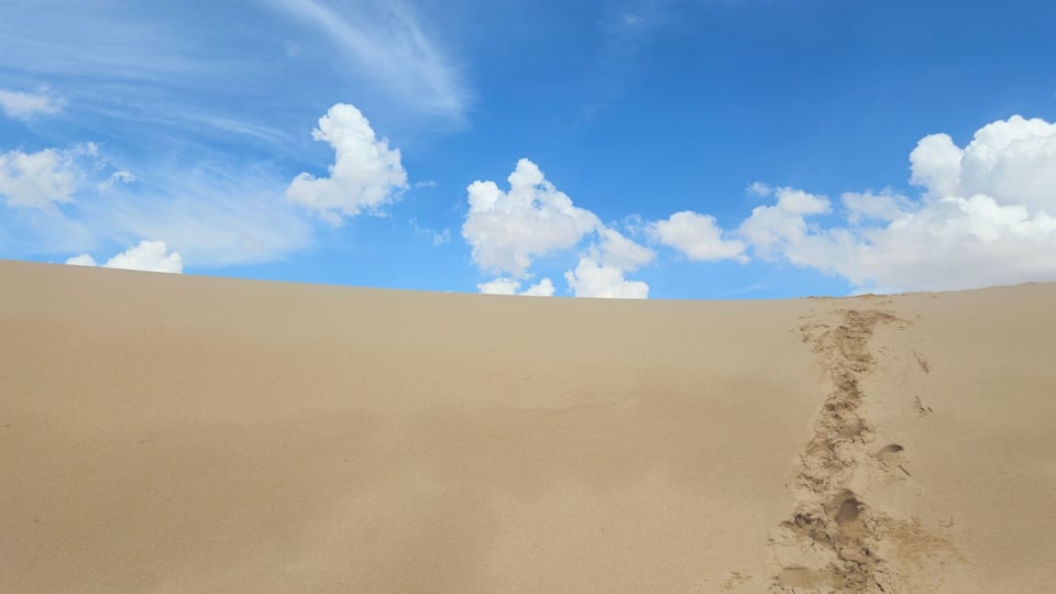 A vast sandy desert with a clear blue sky and scattered clouds.