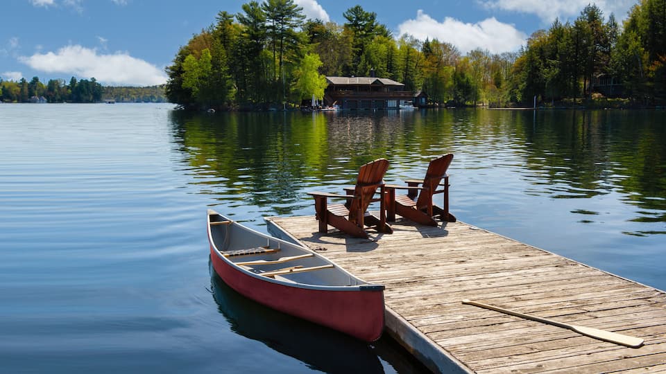 A calm lake with a wooden dock, two Adirondack chairs, and a red canoe.