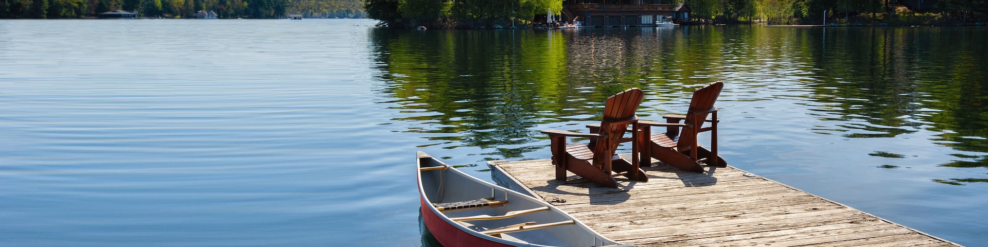 Two Adirondack chairs on a wooden dock overlook the tranquil blue waters of a Muskoka lake in Ontario, Canada. Nearby, a red canoe is tied to the pier, with life jackets and a paddle.