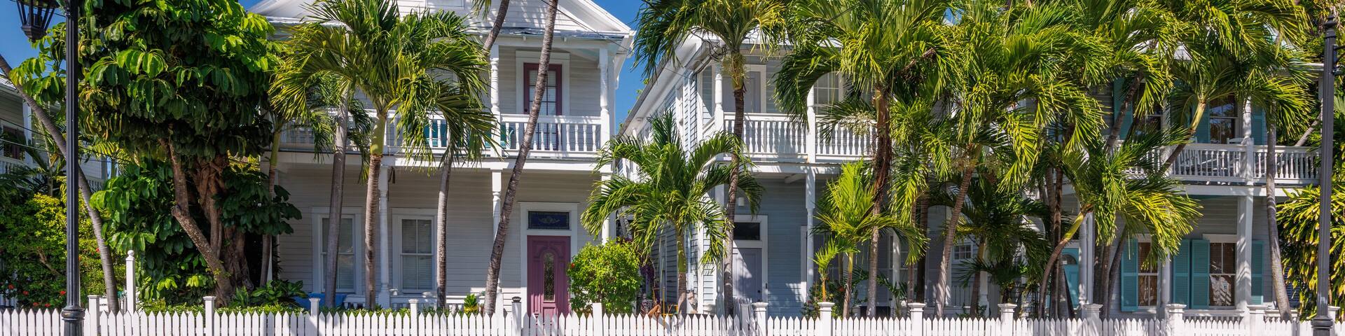 A white colonial-style building with a white picket fence, surrounded by palm trees.