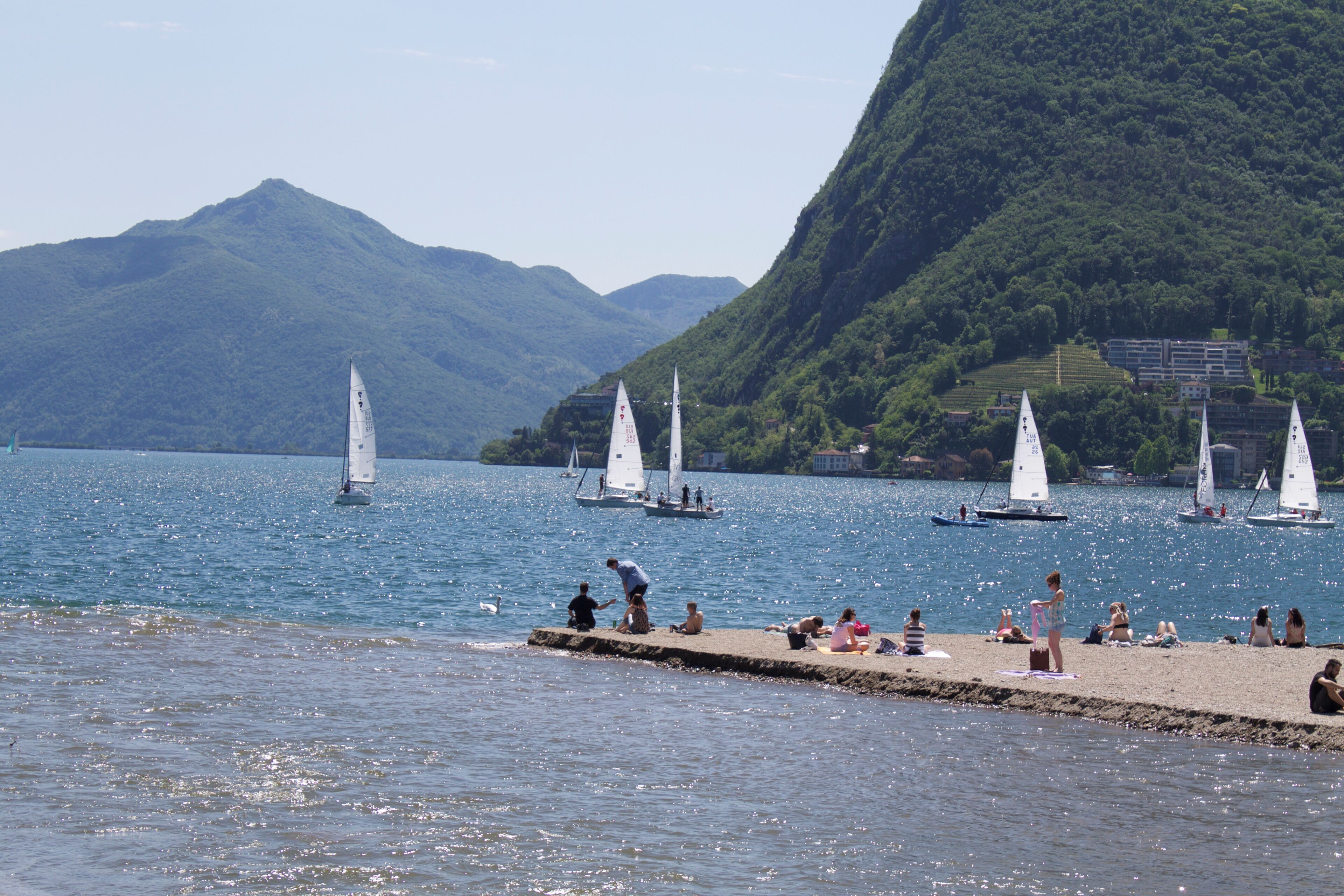 A lake with sailboats, a dock with people sunbathing, and mountains in the background.