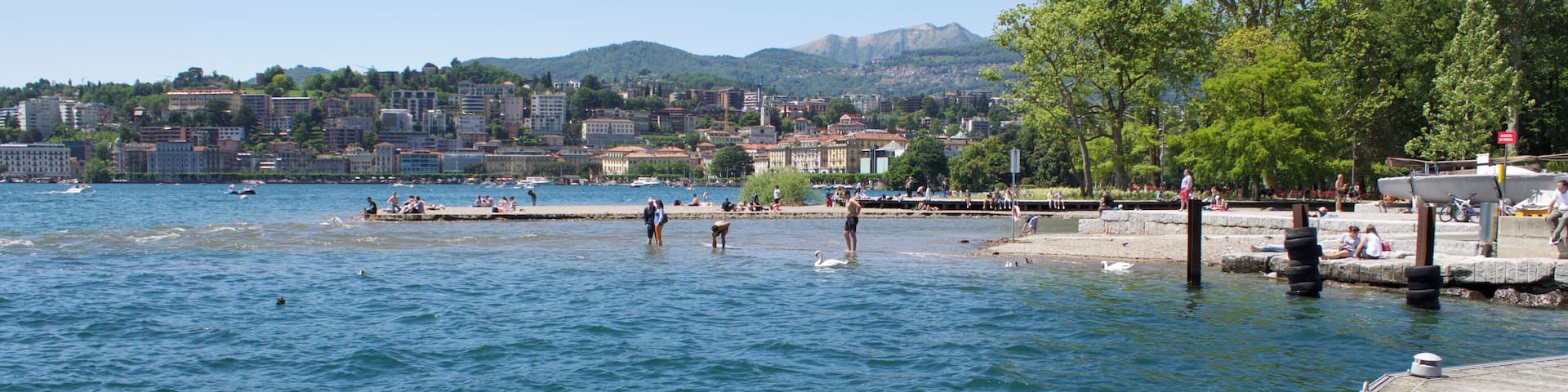 A waterfront with a wooden pier, calm water, and a cityscape with buildings and mountains in the background.