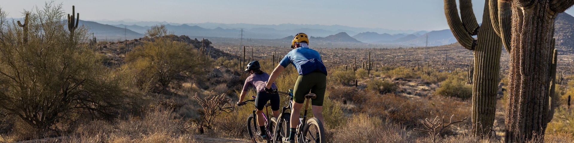 Two cyclists on a dirt trail in a desert landscape with cacti and sparse vegetation.