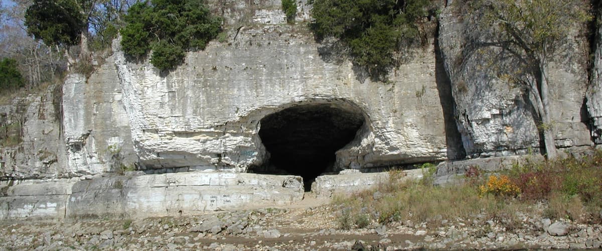A large cave opening in a rocky cliff with sparse vegetation around.